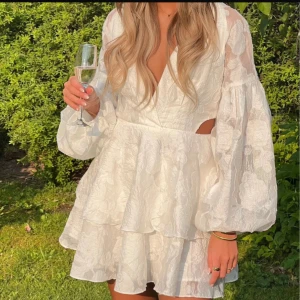 Vit blommig kortklänning Showpo - Superfin vit kortklänning från Showpo med blommigt mönster, v-ringning och volangkjol. Klänningen har ballongärmar, cut out-detalj i midjan och omlottliv. 