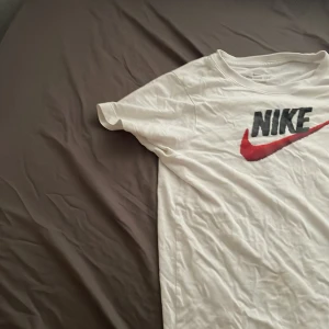 Vit Nike t-shirt med röd swoosh - Vit t-shirt från Nike med klassisk svart logga och röd swoosh framtill. Enkel och stilren design med rund hals och korta ärmar. Perfekt för dig som gillar sportiga och avslappnade outfits.