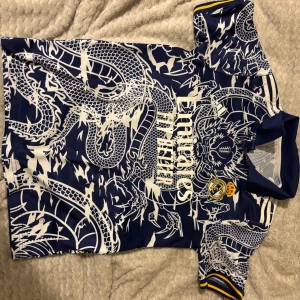 Real Madrid x Adidas blå draktröja - Snygg Real Madrid fotbollströja från Adidas i mörkblått med vitt drakmönster över hela tröjan. Klassisk krage, korta ärmar med gula detaljer och broderat klubbmärke på bröstet. Tillverkad i lätt och ventilerande polyester – perfekt för match eller träning.