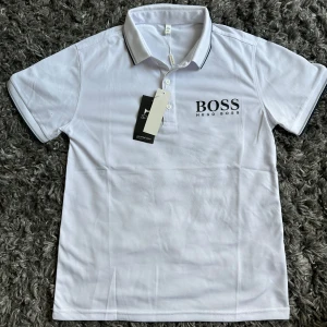 Vit Hugo Boss pikétröja S - Vit pikétröja från Hugo Boss med svart logga på bröstet. Klassisk krage med svarta detaljer och knäppning framtill. Kortärmad modell i bomull, perfekt för en clean och stilren look.