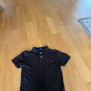 Svart pikétröja från Polo Ralph Lauren - Klassisk svart pikétröja från Polo Ralph Lauren med röd broderad logga på bröstet. Tröjan har korta ärmar, krage och två knappar framtill. Tillverkad i mjuk bomull för en skön känsla och snygg passform.