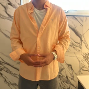 Persikofärgad Oxfordskjorta GANT XL - Snygg persikofärgad Oxfordskjorta från GANT i slim fit. Skjortan har klassisk krage, långa ärmar och knappar framtill. Broderad GANT-logga på bröstet ger en clean och stilren look. Perfekt för dig som gillar preppy stil och vill sticka ut med färg.