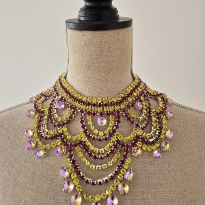 Statement halsband med kristaller - Superfint statement halsband med flera rader av gnistrande gula och vinröda stenar. Halsbandet har stora, lila skimrande kristaller som hänger längs kanten och ger en riktigt lyxig och unik look. Perfekt för dig som vill sticka ut och glänsa extra mycket.