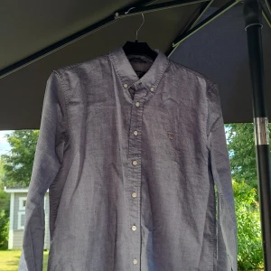 Grå skjorta från Gant, 158/164 - Snygg grå skjorta från Gant med klassisk button down-krage och broderad logga på bröstet. Skjortan har långa ärmar och knäppning framtill. Perfekt för dig som gillar stilrena och tidlösa plagg. Tillverkad i mjuk bomull för skön känsla.