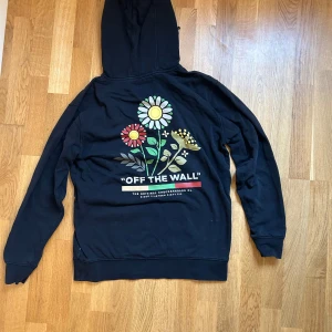 Svart hoodie från Vans med blommor - Svart hoodie från Vans med färgglada blommor och texten 'OFF THE WALL' på ryggen. Liten blom- och Vans-logga på bröstet. Klassisk känguruficka, huva med snörning och långa ärmar. Perfekt för dig som gillar streetstyle och vill sticka ut lite extra.