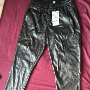 Svarta cargopants i skinnimitation - Snygga svarta cargopants i skinnimitation med elastisk midja och snörning. Byxorna har stora fickor på sidorna och en modern, lite loose passform. Perfekta för dig som vill sticka ut med en edgy stil.