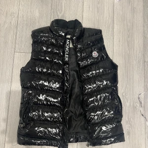 Svart dunväst från Moncler, S - Snygg svart dunväst från Moncler med glansig finish och hög krage. Västen har dragkedja framtill, två fickor med dragkedja och klassisk Moncler-logga på bröstet. Perfekt för lager-på-lager och streetstyle.