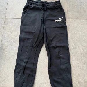 Svarta mjukisbyxor från Puma  - Svarta mjukisbyxor från Puma med vit logga på vänster lår. Byxorna har resår i midjan, dragsko och sidofickor. Tillverkade i mjukt bomullsmaterial, perfekta för chill eller träning. Klassisk och enkel design som funkar till det mesta.