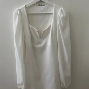 Vit långärmad klänning från NLY One - Stilren vit kortklänning från NLY One med långa puffärmar och djup v-ringning framtill. Klänningen har en rak passform och är tillverkad i ett mjukt, lätt material som ger en elegant känsla. Perfekt för dig som gillar minimalistisk och trendig stil.