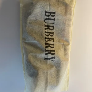 Burberry rutig axelväska med logga - Säljer en klassisk axelväska från Burberry med ikoniskt rutigt mönster i beige, svart, vitt och rött. Väskan har justerbar svart axelrem, detaljer med Burberry-logga och är tillverkad i slitstark polyester. Perfekt storlek för det viktigaste och snygg att bära crossbody.