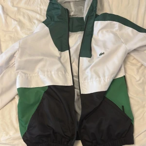 Lacoste vindjacka i grönt, vitt och svart - Snygg vindjacka från Lacoste med coola färgblock i grönt, vitt och svart. Jackan har huva, meshfoder och dragkedja framtill. Ikoniska krokodilloggan på bröstet och elastiska muddar. Perfekt för dig som gillar sportig och stilren vibe.