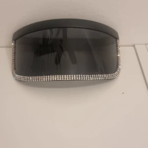 Svarta solglasögon med strasskant - Stora, svarta solglasögon med en bred, rektangulär form och mörka glas. Runt hela bågen sitter en glittrig rad av strass som ger en extra lyxig och trendig känsla. Perfekta för dig som vill sticka ut och addera lite bling till din look.