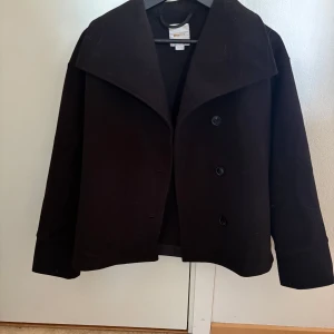 Svart kappa  från Gina Tricot - Stilren svart kappa från Gina Tricot med bred krage och knäppning framtill. Jackan har en klassisk rak passform och långa ärmar, perfekt för dig som gillar en enkel men snygg look. Passar till många olika outfits och är ett måste i garderoben. Storlek 158/164 men motsvarar S dam