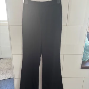Svarta kostymbyxor med bootcut - Snygga svarta kostymbyxor med bootcut-silhuett och klassisk midja. Byxorna har hällor för bälte och stängs med dragkedja och knapp. Perfekta för en clean och stilren look. Köparen står för frakten