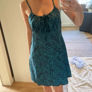 Turkos kortklänning med paisleymönster - Säljer en turkos kortklänning med tunna axelband och svart paisleymönster. Klänningen har snörning vid urringningen och är lätt utsvängd nertill. Perfekt för varma dagar och har en luftig känsla💗