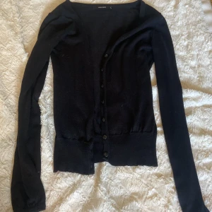 Svart kofta från Vero Moda - En svart, klassisk kofta från Vero Moda i storlek S. Koftan har långa ärmar, v-ringning och knäppning framtill med svarta knappar. Den är stickad och har en enkel design som passar till många olika outfits.