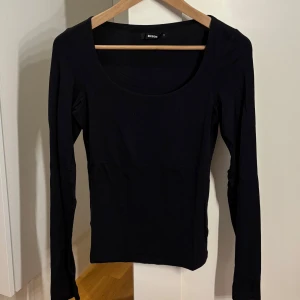 Svart långärmad topp från Bik Bok - En stilren svart långärmad topp från Bik Bok med rundad, djup urringning. Toppen är figurnära och tillverkad i mjukt bomullsmaterial, perfekt att matcha med jeans eller kjol för en clean look. Man kan även bära den bak och fram för att variera outfitsen. 