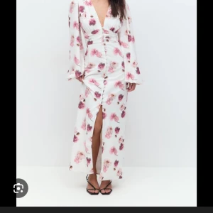 Vit blommig klänning från Gina - Superfin långklänning i vitt med rosa och lila blommor, djup v-ringning och långa ärmar. Klänningen har knappar framtill och en snygg slits. Perfekt för sommarens alla tillfällen. Sitter som en adoore. Blev slutsåld direkt. Använt 1 gång