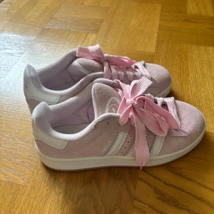 Adidas Campus rosa  - Säljer ett par Adidas Campus sneakers i ljusrosa mocka med vita detaljer och breda rosa skosnören. Klassisk modell med tre ränder på sidan och Adidas-logga på plösen. Skorna har platt sula och rund tå, perfekt för dig som gillar en clean och söt look.