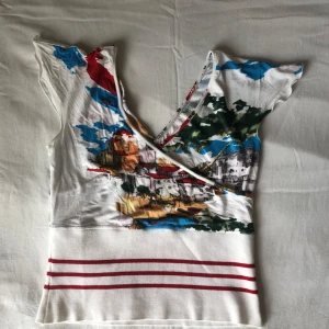 Färgglad omlottopp med tryck - Unik omlottopp i vitt med färgstarkt tryck av stadsmotiv och inslag av blått, rött, grönt och gult. Ser likadan ut på baksidan. Saknar etikett, men gissar på storlek M.