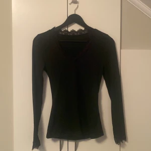 Svart långärmad topp med spetsdetaljer - Säljer en svart långärmad topp med v-ringning och snygga spetsdetaljer vid halsringning och ärmslut. Figurnära passform som ger en stilren look, perfekt att matcha med jeans eller kjol. Enkel men ändå elegant design.