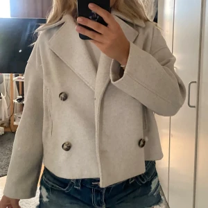 Beige skepparkavaj från Stradivarius - Snygg beige skepparkavaj från Stradivarius med dubbelknäppning och stora knappar i sköldpaddsmönster. Jackan är croppad med bred krage och raka ärmar, perfekt för dig som vill ha en stilren och trendig look. Materialet är mjukt och känns lyxigt.
