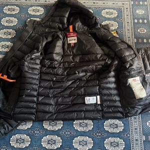 Svart dunjacka Parajumpers med huva - Säljer en svart dunjacka från Parajumpers i storlek M. Jackan har huva, dragkedja framtill, två sidofickor med dragkedja och en patch på ärmen. Klassisk pufferstil med snygga detaljer och orange inslag vid halsen.
