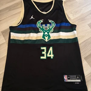 Milwaukee Bucks basketlinne Antetokounmpo - Snyggt Milwaukee Bucks basketlinne med nummer 34 och Antetokounmpo på ryggen. Svart bas med gröna, blå, vita och guldiga ränder över bröstet samt Bucks-logga. Tillverkat i lätt polyester, perfekt för basket eller streetwear.