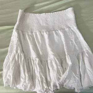 Vit volangkjol med smockmidja - Superfin vit kortkjol med bred smock i  midjan och volangkant nertill. Kjolen är luftig och har en flowy känsla, perfekt för varma dagar. Passar dig som gillar en söt och avslappnad stil.