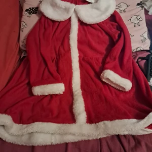 Röd julklänning med fuskpälsdetaljer (barn) - Supermysig röd klänning med vita fuskpälsdetaljer på krage, ärmslut och nederkant. Perfekt för julens alla tillfällen! Klänningen är gjord i mjuk polyester och har lång ärm. Söt och klassisk tomte-look som sticker ut. (Har en liten skada där man stänger bakifrån, men är inte så synlig)