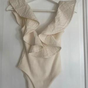 Beige volangbody med djup ringning - Supersnygg beige body med stora volangdetaljer längs axlar och urringning. 