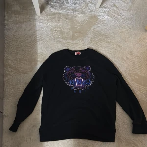 Kenzo tröja - Svart sweatshirt från Kenzo med ikoniskt tigertryck i lila och blå toner på bröstet. Tröjan har rund halsringning och långa ärmar. Perfekt för dig som gillar streetwear och vill sticka ut med en snygg logga framtill.