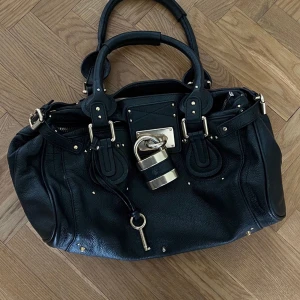 Chloé svart handväska i skinn - Säljer en ikonisk svart handväska från Chloé i äkta skinn med guldiga metalldetaljer och ett stort lås framtill. Väskan har dubbla handtag, dragkedja och nyckel som hänger i en rem. Perfekt för dig som gillar statement-accessoarer.