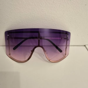Oversized lila visirsolglasögon - Stora, futuristiska solglasögon med visirform och lila till rosa tonade glas. Ramlös design med svarta skalmar och metallbåge runt nederdelen. Perfekt statement-piece för dig som vill sticka ut.