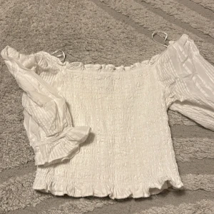 Vit offshoulder blus med volang - Supersöt vit offshoulder blus med smockad kropp och långa puffiga ärmar. Blusen har volangdetaljer vid ärmslut och nederkant, vilket ger en romantisk vibe. Perfekt för dig som gillar en feminin och trendig stil.