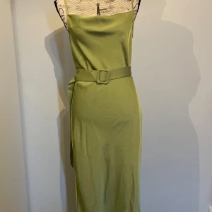 Satin långklänning med bälte - Snygg ljusgrön långklänning i satinliknande material med smala axelband och markerad midja med matchande bälte. Klänningen har en slits vid sidan och en stilren, minimalistisk look som känns både modern och chic. (Finns en liten fläck, se sista bilden)