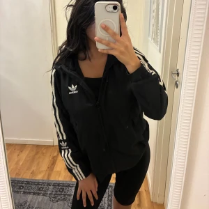 Svart Adidas vindjacka med huva UNISEX - Svart vindjacka från Adidas med klassiska vita ränder längs ärmarna och broderad logga på bröstet och ena ärmen. Jackan har huva, dragkedja framtill och två fickor. Perfekt för en sportig och chill stil. Aldrig kommit till användning. 