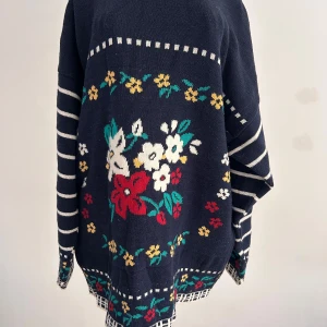 Blommig tätstickad tröja från Lindex - Mörkblå tätstickad tröja från Lindex med blommigt mönster framtill. Ärmarna är randiga i vitt och blått. Perfekt för dig som gillar retro vibes och färgglada detaljer.