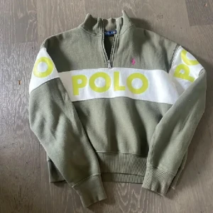 Olivgrön half zip tröja från Polo Ralph Lauren - Olivgrön långärmad tröja från Polo Ralph Lauren med vit och gul POLO-logga över bröst och ärmar. Tröjan har half zip, ribbade muddar och en rosa broderad logga på bröstet. Tillverkad i mjuk bomull, perfekt för en chill och sportig look.