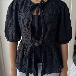 Svart blus med knyte och struktur -  svart blus med puffiga ärmar och snygg struktur i tyget. Blusen har två knyten framtill och en lätt volang nertill🩷