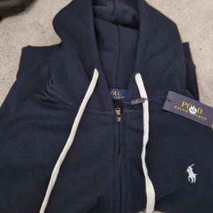 Marinblå hoodie från Polo Ralph Lauren - (En helt ny och oanvänd ralph lauren zip tie / hoodie) ( Det är en M och är för liten för mig, Men det är en mindre M och en person i S hade passat bra i den )(kom dms för att diskutera pris) snygg marinblå hoodie från Polo Ralph Lauren med vit dragsko och dragkedja framtill. Klassisk logga broderad på bröstet.