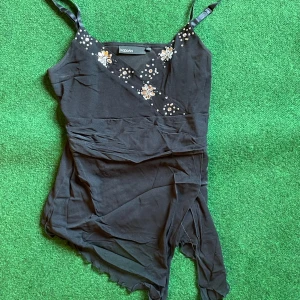 Svart topp med pärlor KappAhl - Svart linne från KappAhl med omlott-look och smala axelband. Toppen har dekorativa pärlor och blommiga broderier framtill, vilket ger en extra snygg detalj. Perfekt för dig som gillar unika toppar med lite extra bling.