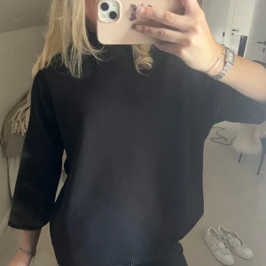 Svart polotröja med trekvartsärm - En svart polotröja med hög krage och trekvartsärmar. Tröjan har en loose passform och är enkel och stilren, perfekt att matcha med jeans eller kjol. Materialet känns mjukt och bekvämt mot huden.