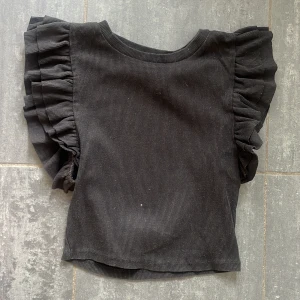 Svart volangtopp från Zara - Supersnygg svart topp från Zara med breda volanger längs axlarna. Toppen är ribbad och har en rak, enkel passform som känns både trendig och cool. 