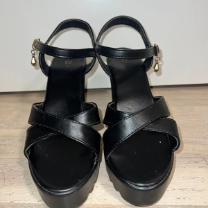 Svarta sandaler med klack och platå - Snygga svarta sandaler med bred klack och platåsula. Remmarna är i skinnimitation och går i kors över foten. Runt vristen sitter en smal rem med spänne och en liten hängande pärldetalj i metall. Perfekta för dig som vill ha lite extra höjd och stil.