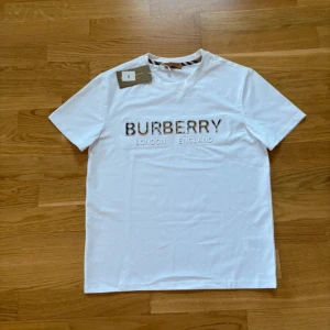 Vit Burberry t-shirt med logga - Vit t-shirt från Burberry med klassisk rund hals och korta ärmar. Stor Burberry-logga framtill med rutiga detaljer i bokstäverna. Tillverkad i mjuk bomull för en clean och lyxig känsla. Perfekt för dig som gillar stilrena statement-plagg.