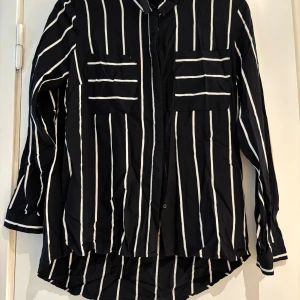 Svartvit randig skjorta från ONLY - Svart skjorta från ONLY i storlek 38 med vita vertikala och horisontella ränder. Skjortan har två bröstfickor, klassisk krage och knäppning framtill. Långärmad modell med avslappnad passform, perfekt för en cool och stilren look.