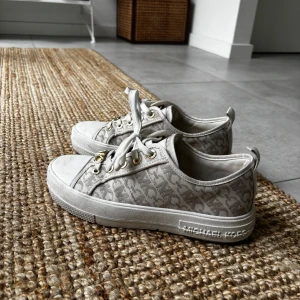 Vita sneakers från Michael Kors - Snygga vita sneakers från Michael Kors med diskret MK-mönster och guldfärgade detaljer. Skorna har snörning, rund tå och platt sula. Tillverkade i textil och syntet med logga på sidan och plösen. Perfekta för en trendig och avslappnad look.