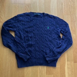 Marinblå kabelstickad tröja Ralph Lauren - Kabelstickad marinblå tröja från Ralph Lauren med rund halsringning och grönt broderat logomärke på bröstet. Tröjan har långa ärmar och ribbade muddar vid ärmslut och nederkant. Perfekt för lager-på-lager och snygg till jeans.