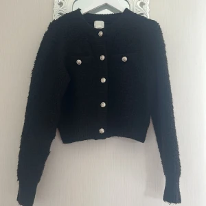 Svart kofta med silverknappar S  - Snygg svart kofta med silvriga dekorativa knappar framtill och på fickorna. Koftan är stickad med en fluffig struktur, har rund halsringning och långa ärmar. Perfekt för dig som gillar en klassisk men trendig look.